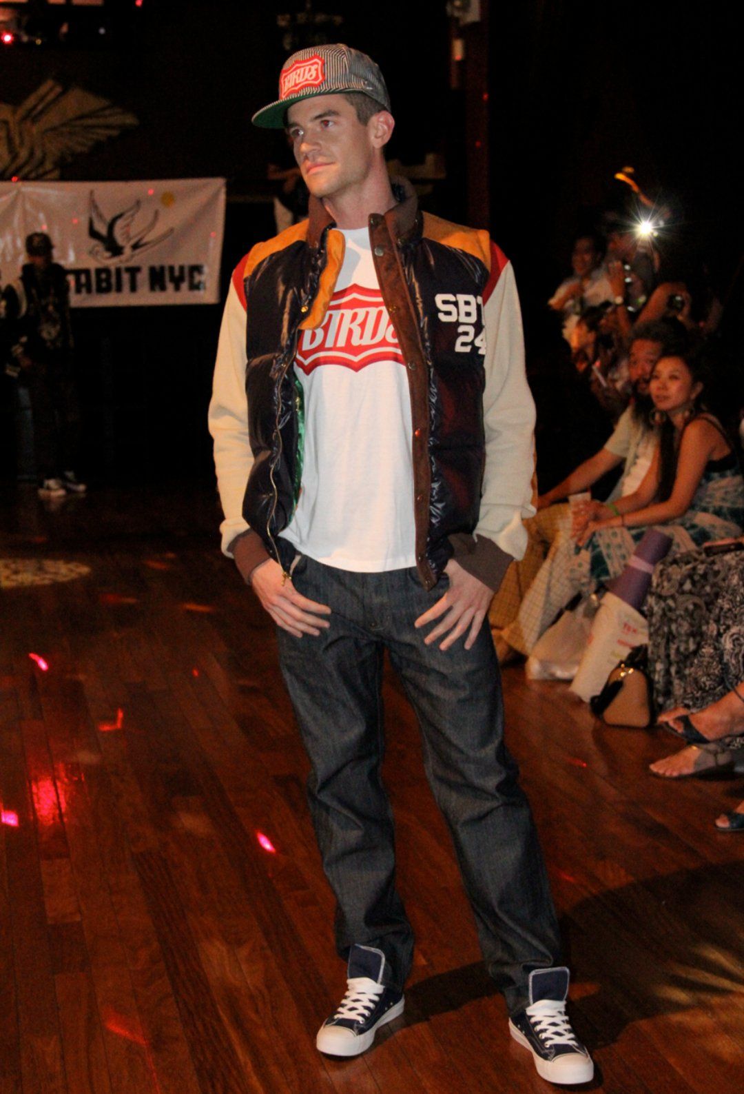 Male fit model Joe Cummings on a New York City runway wearing a streetwear puffer vest, graphic tee, jeans and cap